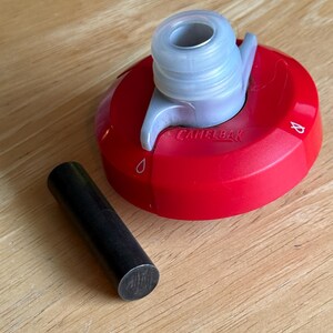 May include: A red CamelBak water bottle cap with a translucent gray spout and a small black cylindrical pin. The cap features a rotating mechanism with water drop icons and the brand name CamelBak embossed on the top surface of the red plastic.