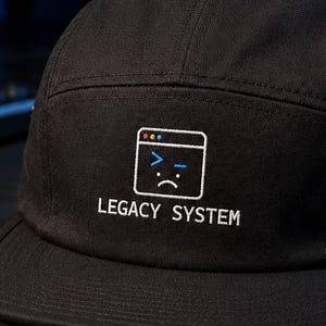 May include: A black five-panel cap featuring an embroidered graphic of a computer window icon with a sad face and the text LEGACY SYSTEM in white. The hat has a flat brim and a structured crown with a small metal eyelet on the side panel.