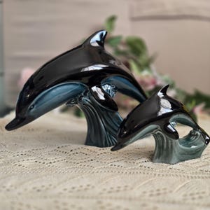 Könnte beinhalten: Zwei glänzende Keramik-Delfinfiguren in einem dunklen Schwarz und gedämpften Blau. Jeder Delfin ist so dargestellt, als würde er aus stilisierten, texturierten Wellen springen. Der größere Delfin steht links, ein kleinerer rechts daneben.