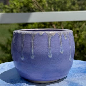May include: A rounded ceramic planter with a smooth lavender purple glaze. The upper rim features a decorative white drip pattern that cascades down the sides of the vessel, creating a contrast against the solid purple body of the container.