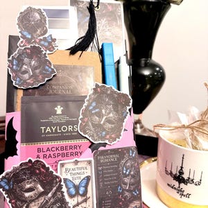 May include: A gift set featuring a brown journal, a Taylors of Harrogate Blackberry & Raspberry tea bag, and various stickers depicting masquerade masks and blue butterflies. A white mug with a gold rim and the text midnight spell sits nearby.