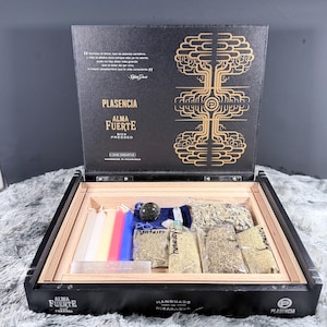 May include: A black wooden box with a hinged lid featuring gold tree graphics and text including Plasencia Alma Fuerte. Inside are five colored taper candles, a dark stone, a clear crystal, and four clear plastic bags of dried herbs labeled Plantain, Harmony, and Rosemary.