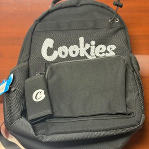 May include: Black backpack featuring the word "Cookies" in white. It includes a front pocket and a small wallet with a "C" logo. The backpack has adjustable straps and a top handle. The bag is made of a durable material.