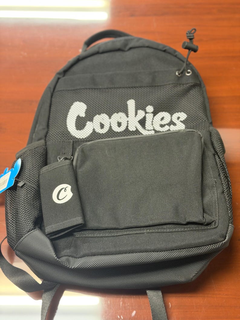 May include: Black backpack featuring the word "Cookies" in white. It includes a front pocket and a small wallet with a "C" logo. The backpack has adjustable straps and a top handle. The bag is made of a durable material.