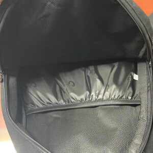 May include: An open black backpack, showing the interior. The main compartment and a smaller pocket are visible. The interior fabric has a textured pattern. The zip is partially open, revealing the inside of the bag.