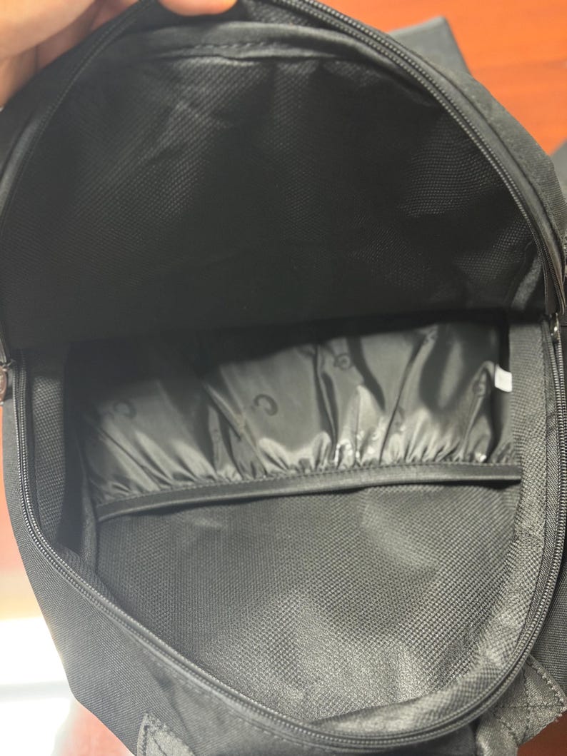 May include: An open black backpack, showing the interior. The main compartment and a smaller pocket are visible. The interior fabric has a textured pattern. The zip is partially open, revealing the inside of the bag.