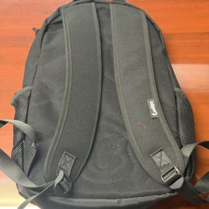 May include: Black backpack with padded shoulder straps and a top handle. The backpack features a stitched design on the back and the word "Cookies" printed on the side. The backpack has mesh side pockets and adjustable straps.