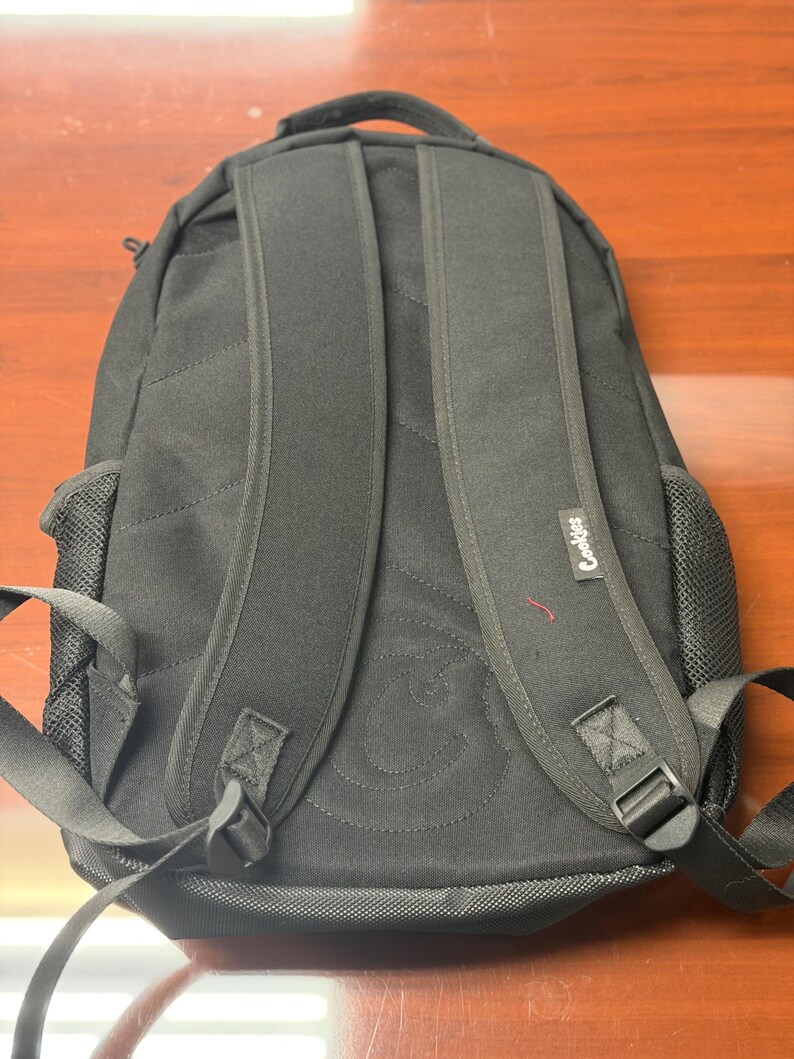May include: Black backpack with padded shoulder straps and a top handle. The backpack features a stitched design on the back and the word "Cookies" printed on the side. The backpack has mesh side pockets and adjustable straps.