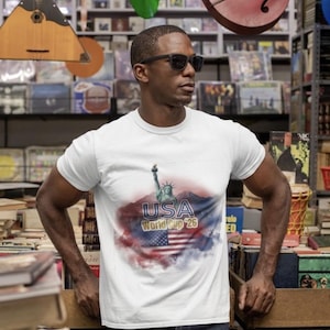 Pode incluir: Uma t-shirt branca de manga curta com uma impressão gráfica da Estátua da Liberdade, o texto USA World Cup '26 e uma bandeira americana sobre um fundo de nuvens em estilo aguarela vermelho, branco e azul no peito.