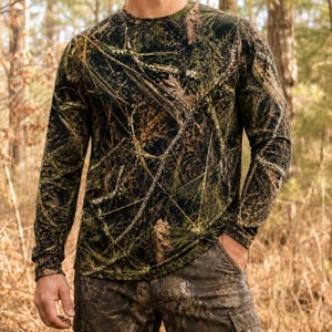 May include: A long-sleeve crew neck shirt featuring a realistic forest floor camouflage pattern in shades of dark brown, black, and forest green, depicting pine needles, twigs, and fallen leaves across the entire garment surface.
