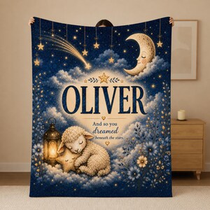 May include: A dark blue fleece blanket featuring a sleeping lamb hugging a star next to a glowing lantern. The design includes a crescent moon, stars, a shooting star, and blue flowers. The text reads OLIVER And so you dreamed beneath the stars.