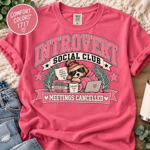 Puede incluir: Una camiseta rosa coral con un gráfico de un perro con gafas de sol, envuelto en una manta, con libros, un portátil y una taza. El texto dice INTROVERT SOCIAL CLUB y MEETINGS CANCELLED, con lomos de libros etiquetados como GOOD BOOKS, COMFY BLANKETS, PEACE & QUIET, HAPPY ME.