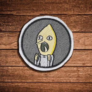 May include: A circular embroidered patch featuring a yellow lemon-shaped character with a wide-open mouth, dark eyes, and a pointed nose. The character wears a white shirt and is set against a dark gray background with a thick white border.