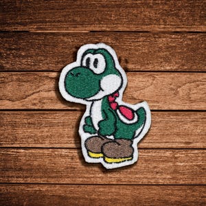 May include: An embroidered patch featuring a green dinosaur character with a white belly, large white eyes, a red saddle on its back, and brown shoes with yellow soles, outlined in a white border against a dark brown wooden background.