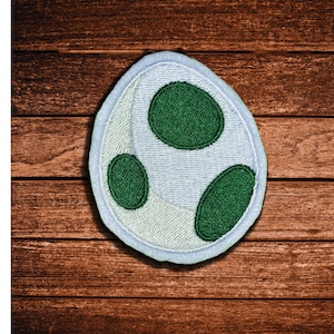 May include: An oval-shaped embroidered patch featuring a white background with three distinct dark green oval spots. The patch has a light blue stitched border and a textured surface created by dense thread work throughout the entire design.
