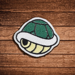 May include: An embroidered patch featuring a stylized turtle shell design. The shell has a dark green top section with black outlines, a thick white middle border, and a pale yellow bottom section with black curved lines, all finished with a white edge.
