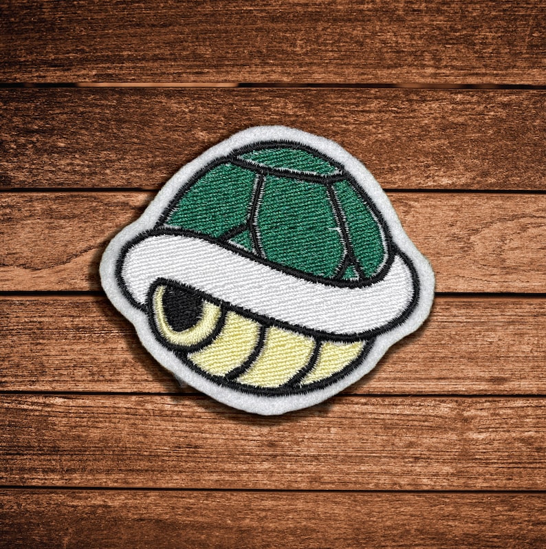 May include: An embroidered patch featuring a stylized turtle shell design. The shell has a dark green top section with black outlines, a thick white middle border, and a pale yellow bottom section with black curved lines, all finished with a white edge.