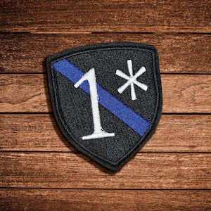 Könnte beinhalten: Ein schildförmiger, bestickter Aufnäher mit schwarzem Hintergrund, der einen diagonalen blauen Streifen, eine große weiße Ziffer 1 und ein weißes Sternchen-Symbol aufweist. Der Aufnäher hat einen schwarzen gestickten Rand zur Befestigung an Textilien.