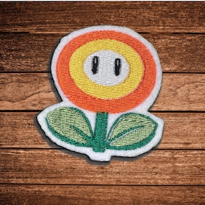 May include: An embroidered patch shaped like a flower with a white center, two black vertical eyes, a yellow ring, and an orange outer border. The flower has a green stem and two green leaves, all outlined in white thread on a white fabric base.