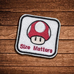 May include: A square embroidered patch with a white background and a black border. The center features a red and white mushroom graphic above the red embroidered text "Size Matters" in a simple, bold, sans-serif font style.