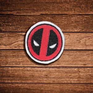 Puede incluir: Un parche bordado circular con un diseño en rojo y negro, una franja vertical roja central y dos detalles blancos en forma de ojos. El parche tiene un borde blanco grueso y un acabado de hilo texturizado en toda su superficie.