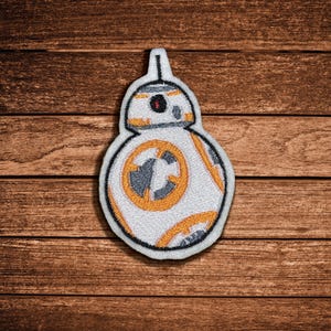 May include: A white, orange, and gray embroidered patch shaped like a spherical robot. The design features a circular body with orange and gray geometric patterns and a smaller dome-shaped head with a single dark eye and a small antenna on top.