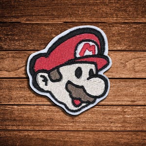 May include: An embroidered patch featuring the head of a character wearing a red cap with a white letter M. The design includes a beige face, a large mustache, and black outlines, all set against a white border on a dark wooden background.