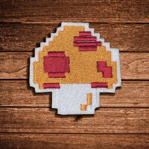 May include: A pixelated mushroom patch featuring a golden yellow cap with dark red square accents and a white stem. The design is rendered in a blocky, digital style with a white border surrounding the entire embroidered mushroom shape.