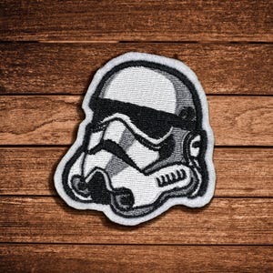 May include: An embroidered patch featuring a black and white Stormtrooper helmet design. The patch has a white border and detailed stitching that outlines the iconic helmet shape, including the black eye lenses and the distinct facial features.
