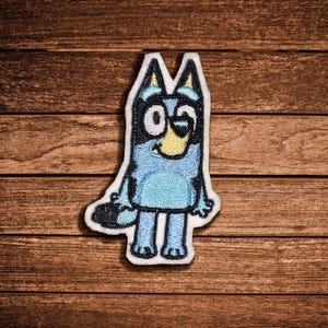 May include: An embroidered patch featuring a cartoon blue dog character with upright ears, a light blue body, a yellow muzzle, and a black nose. The character has large white eyes with black pupils and a dark blue tail, all outlined in black thread.