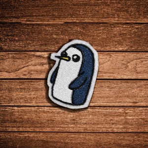 May include: An embroidered patch featuring a stylized penguin with a white chest, dark blue back, and a small yellow beak. The design is outlined in white thread, creating a clean border around the penguin figure, which is depicted in a simple, graphic style.