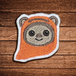 May include: An embroidered patch featuring a stylised creature face with large black eyes and a small nose, framed by a bright orange hood with a white border. The design includes rounded ears and a soft beige face area with detailed stitching.