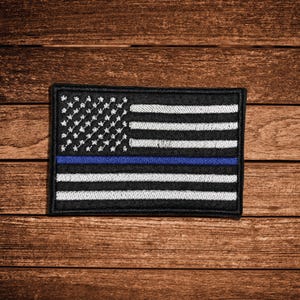 May include: A rectangular embroidered patch featuring a stylized American flag design. The patch is primarily black with white embroidered stars and stripes, and a single horizontal blue stripe running through the center of the flag design.