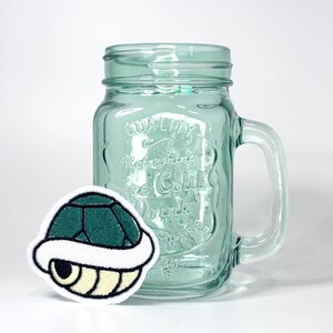 May include: A light green glass mason jar with a handle and embossed text reading Quality Refreshing Ice Cold Drink, accompanied by a circular embroidered patch featuring a dark green and cream colored turtle shell graphic.