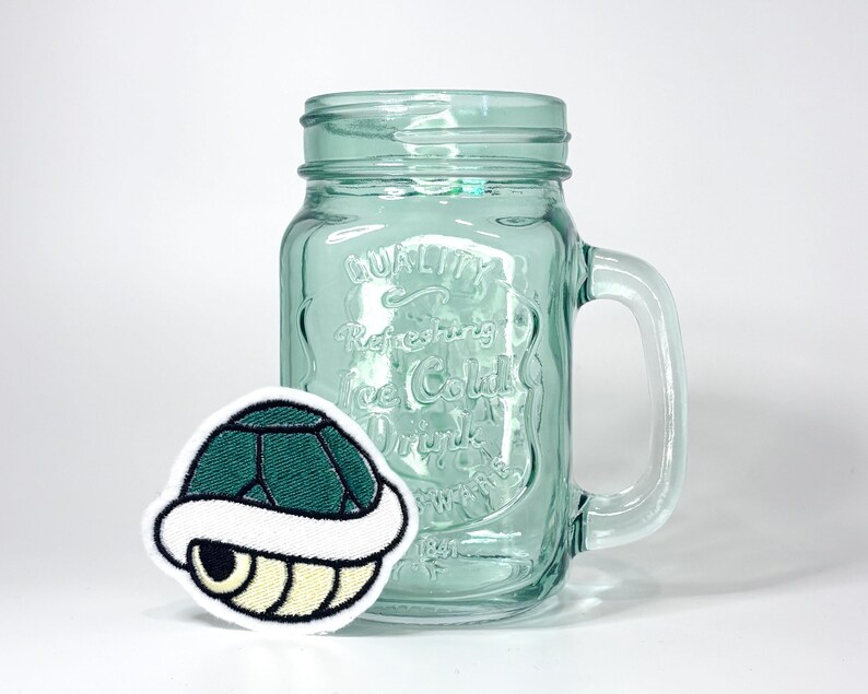 May include: A light green glass mason jar with a handle and embossed text reading Quality Refreshing Ice Cold Drink, accompanied by a circular embroidered patch featuring a dark green and cream colored turtle shell graphic.