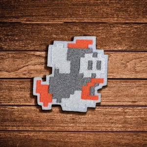 May include: A pixel art style embroidered patch featuring a stylized character design. The figure is composed of grey, white, and orange thread, creating a blocky, retro-inspired aesthetic with distinct geometric shapes and a jagged, stepped outline.