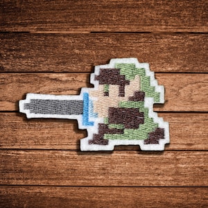 Può includere: Una toppa ricamata raffigurante un personaggio pixelato di profilo, nei toni del verde, marrone e beige, che impugna una lunga spada grigia orizzontale con un'elsa blu brillante, il tutto bordato da un spesso contorno bianco su legno.