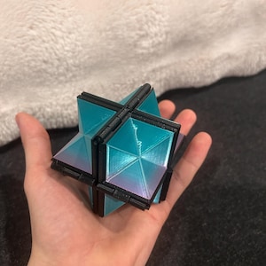 Pode incluir: Um brinquedo de quebra-cabeça geométrico em forma de estrela com molduras de plástico preto e painéis triangulares iridescentes em tons de azul-petróleo e roxo. O objeto é construído com vários segmentos articulados que permitem a sua dobra.