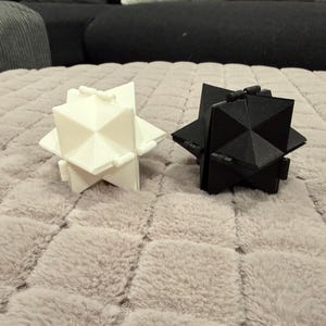 Pode incluir: Dois objetos geométricos em forma de estrela, um em branco sólido e outro em preto sólido. Cada objeto apresenta múltiplas faces triangulares pontiagudas e pequenos conectores tipo dobradiça ao longo das bordas, criando uma estrutura complexa.