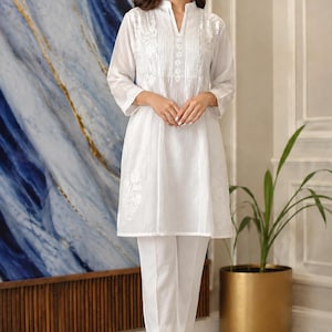 May include: A white tunic featuring a V-neckline with vertical pleats and intricate white floral embroidery on the chest and sleeves, paired with matching white straight-leg trousers that have delicate embroidery along the bottom hem.