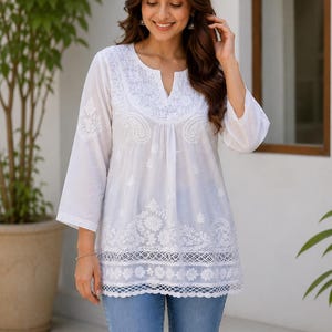 May include: A white tunic featuring intricate white floral embroidery and paisley patterns across the chest and hem. The garment has three-quarter length sleeves, a notched neckline, and a scalloped lace border along the bottom edge.
