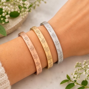 May include: Three metal bangle bracelets in rose gold, yellow gold, and silver tones. Each bracelet features a continuous engraved pattern of small flowers and leaves along the outer surface. The bangles are worn together on a wrist.