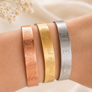 May include: Three metal cuff bracelets in copper, gold, and silver tones. The copper bracelet is engraved with the text NO FLOWERS. The gold bracelet features cursive script text. The silver bracelet displays the word RISE and a sun icon.
