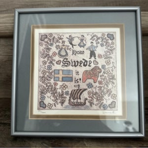 May include: A framed print featuring a folk art illustration with the text "how Swede it is!". The design includes a Swedish flag, a Dala horse, a Viking ship, and figures in traditional clothing, all surrounded by a floral border in a silver frame.