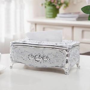 May include: A silver tissue box with intricate floral patterns. The rectangular box features a hinged lid and rests on four curved feet. The design includes raised rose motifs and decorative trim, with white tissues protruding from the top.