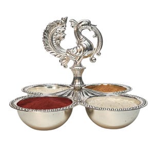 May include: A silver-colored spice server with four round bowls. The server is topped with a decorative peacock design. The bowls contain red, brown, and white spices. The server is designed to hold and display spices.