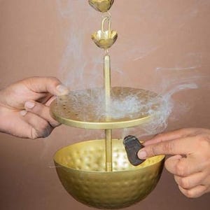 May include: A gold-coloured incense burner with a bowl-shaped base and a perforated disc above. The burner is suspended from a chain of decorative flower-shaped elements. Smoke rises from the incense, creating a hazy effect.