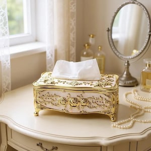 May include: A decorative tissue box with a gold floral design and a cream base. The rectangular box has rounded corners and sits on four small feet. White tissues are visible at the top. A decorative mirror and perfume bottles are in the background.