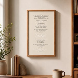 May include: A framed print with a poem in a wooden frame hangs on a wall. The poem is printed in black ink on a cream-colored background. A mug, a notebook, and a pen sit on a wooden desk in front of the print. A vase with greenery is on the left.