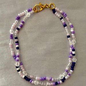 Peut inclure: Un bracelet à deux rangs de perles avec un fermoir mousqueton doré. Le bracelet est composé de petites perles rondes dans des tons de violet clair, violet foncé et blanc, entrecoupées de perles transparentes à facettes effet cristal.